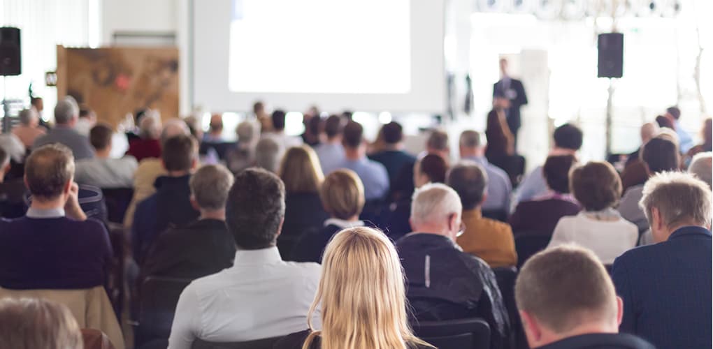 A crowd of seated individuals watch a speaker give a presentation