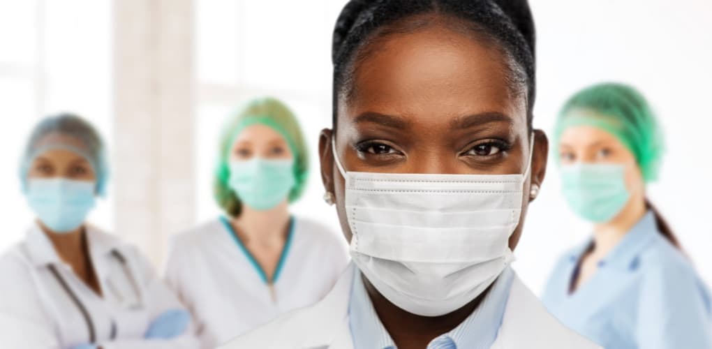 A female physician wearing a medical mask looks into the camera with three other physicians wearing medical masks are in the background.