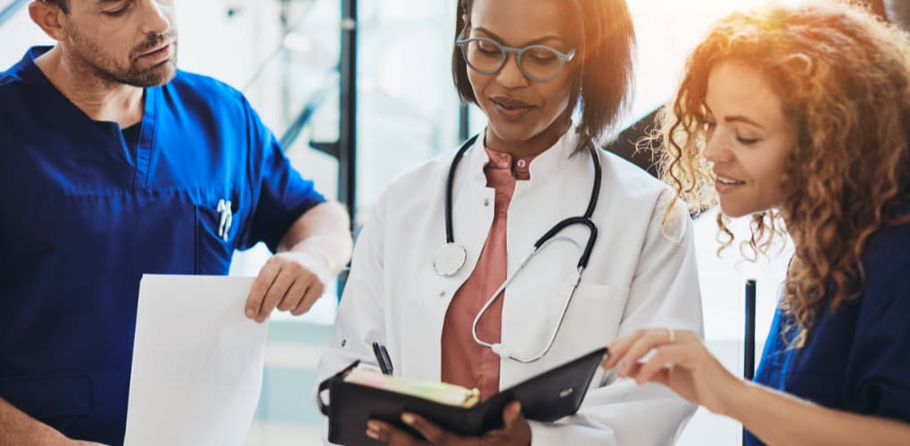 Three doctors review data in a binder. The two doctors on each end have blue scrubs and the doctor holding thee binder in the middle is wearing a white lab coat with a stethoscope around her neck.