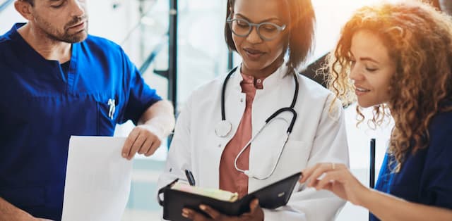 Three doctors review data in a binder. The two doctors on each end have blue scrubs and the doctor holding thee binder in the middle is wearing a white lab coat with a stethoscope around her neck.