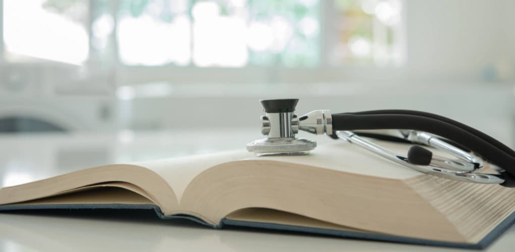 A thick book lays open and face up on a table. A stethoscope sits on top of one side of the book.