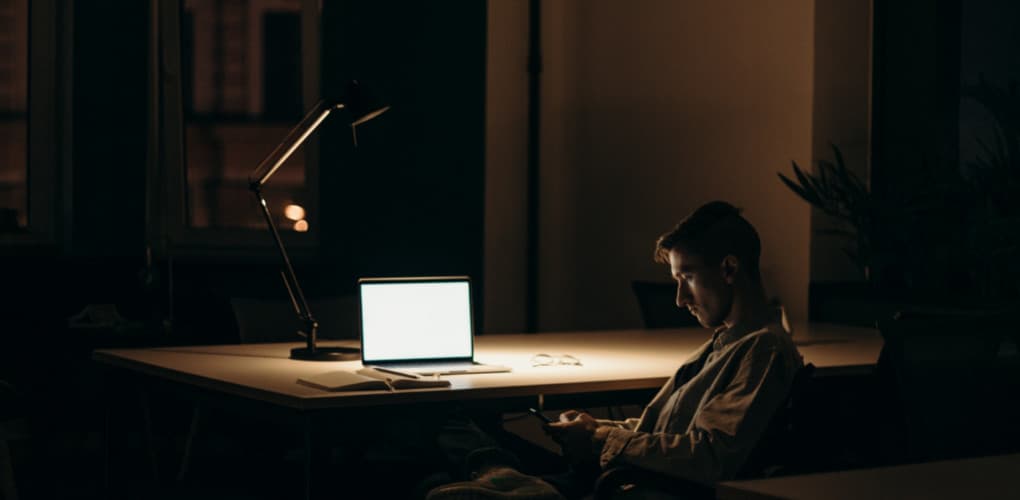 person sitting at desk in a dimly lit room looking at phone with computer open