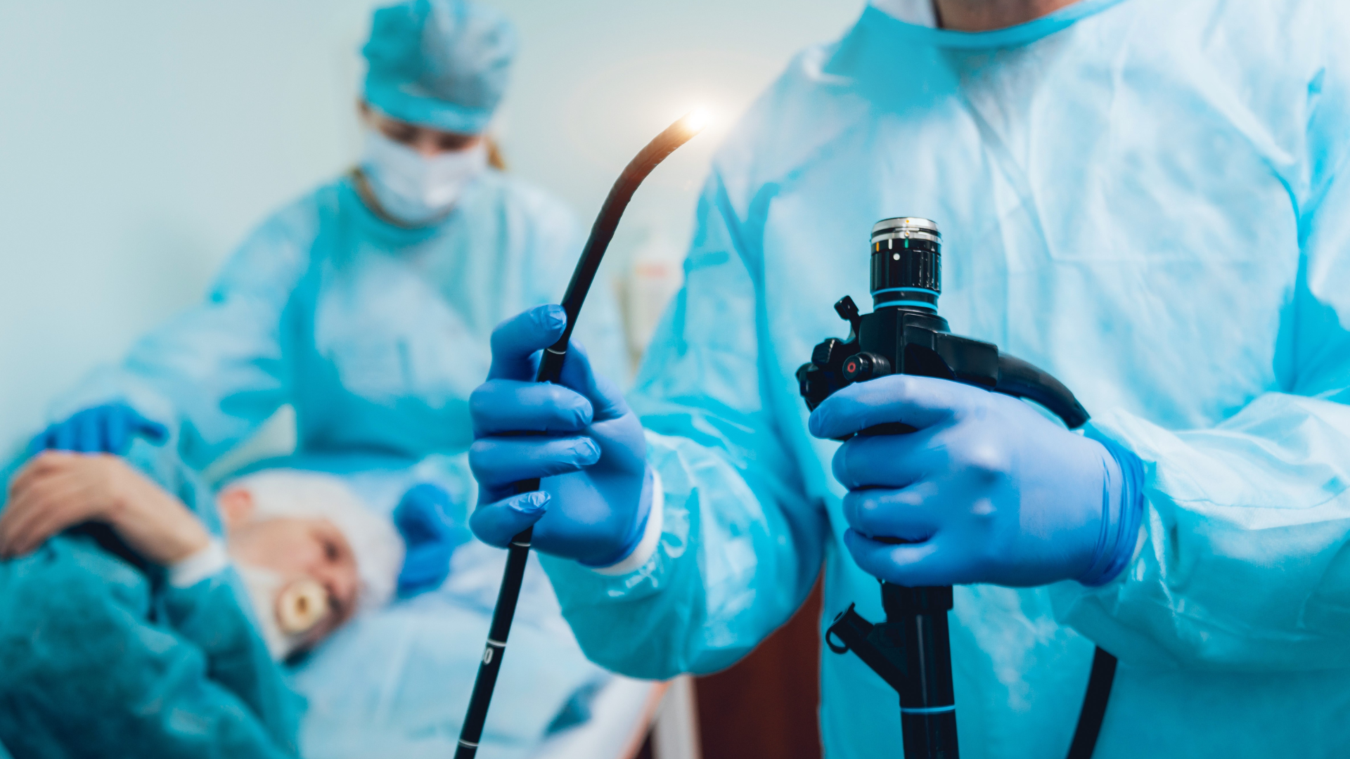 A medical team conducts a procedure with an endoscope. A healthcare professional holds the scope, while a patient lies on a bed in the background, supported by another team member.